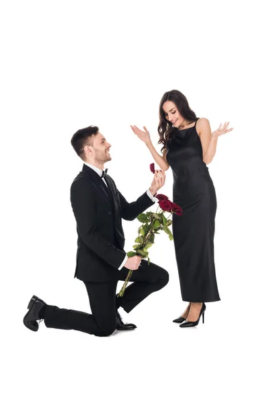 Glücklicher Mann mit roten Rosen, der einem aufgeregten Mädchen, isoliert auf weiß, einen Heiratsantrag macht — Stockfoto