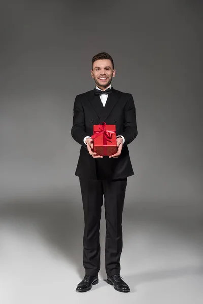 Handsome elegant man in black tuxedo and tie bow gifting red present on grey — Stock Photo