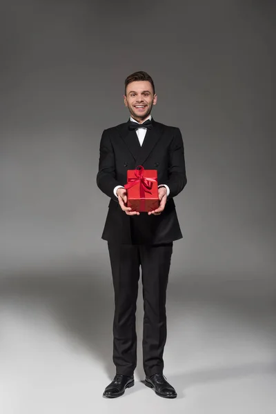 Bonito elegante homem em preto smoking e gravata arco presente vermelho no cinza — Fotografia de Stock