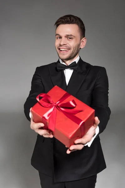 Happy elegant man in black tuxedo and tie bow gifting red present isolated on grey — Stock Photo