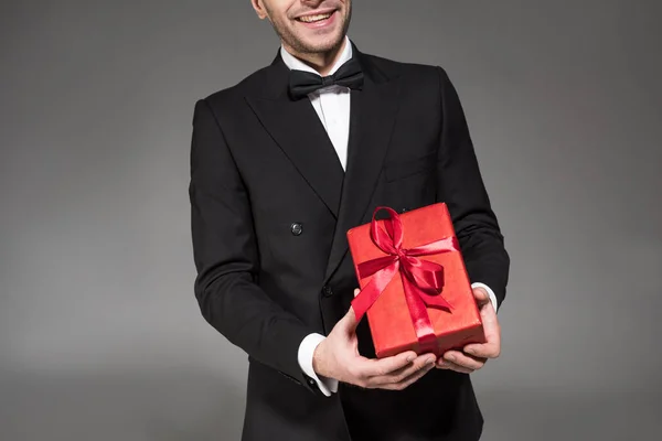 Vista recortada del hombre sonriente en esmoquin negro y corbata de lazo sosteniendo caja de regalo roja aislada en gris - foto de stock