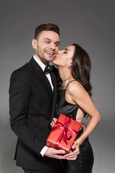 Feliz mulher beijando bonito namorado e segurando caixa de presente no dia dos namorados, isolado no cinza — Fotografia de Stock