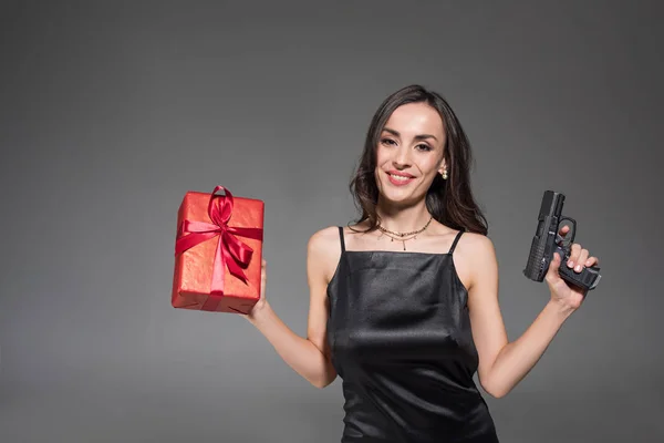 Attractive smiling woman holding red gift box and gun isolated on grey — Stock Photo