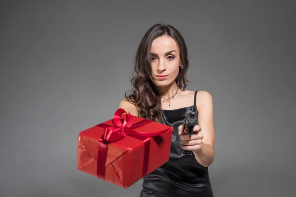 Серьезная брюнетка с красным подарком и пистолетом, изолированным на сером — стоковое фото