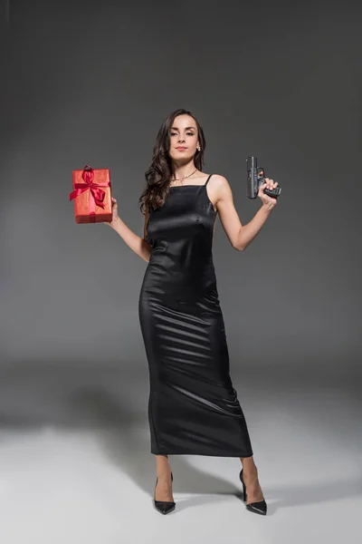 Elegant woman in black dress holding red gift and gun on valentines day isolated on grey — Stock Photo
