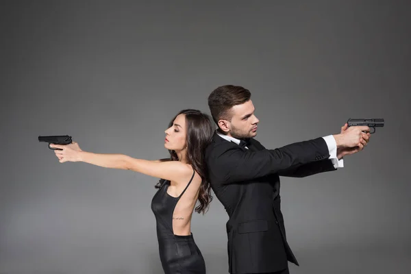 Confiante casal de agentes secretos em roupas pretas visando com arma, isolado em cinza — Fotografia de Stock