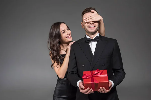 Hermosa mujer cerrando los ojos y haciendo sorpresa a novio con presente en día de San Valentín, aislado en gris - foto de stock