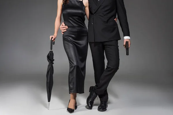 Recortado vista de joven pareja de asesinos en negro ropa posando con arma y paraguas en gris - foto de stock