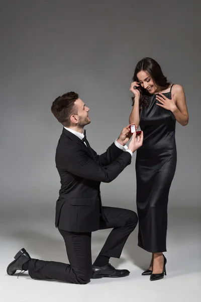Jovem namorado segurando anel de proposta na caixa para namorada atraente no cinza — Fotografia de Stock