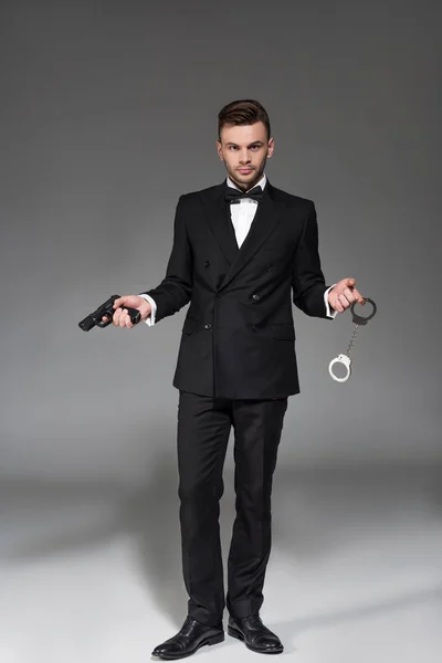 Elegant secret agent in tuxedo holding handgun and handcuffs on grey — Stock Photo