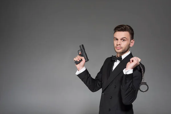 Beau agent secret en costume noir tenant arme de poing et menottes, isolé sur gris — Photo de stock