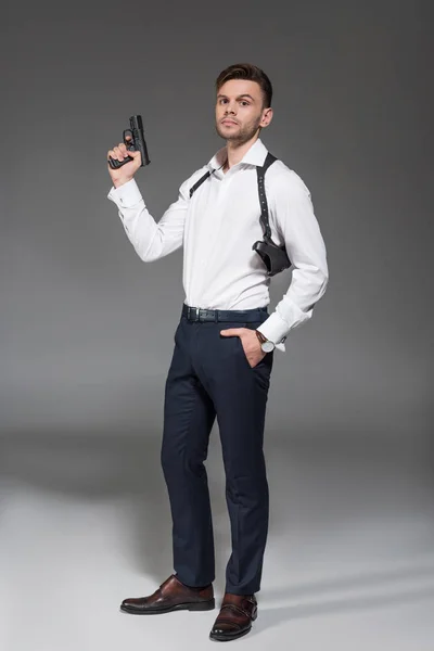 Guapo joven agente secreto con funda sosteniendo pistola en gris - foto de stock