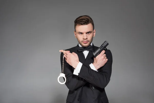 Серйозний секретний агент в чорному смокінгу тримає пістолет і наручники, ізольовані на сірому — стокове фото