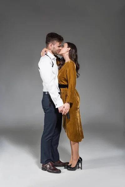Felice bella donna in abito d'oro baciare agente segreto con pistola su grigio — Foto stock