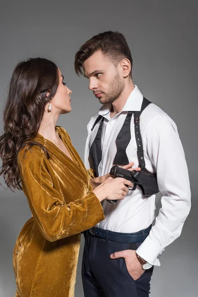 Hermosa mujer en vestido de oro tomando arma de la funda de agente secreto, aislado en gris - foto de stock