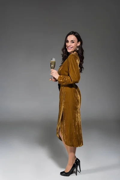 Привлекательная счастливая женщина в золотом платье с бокалом шампанского на сером — стоковое фото