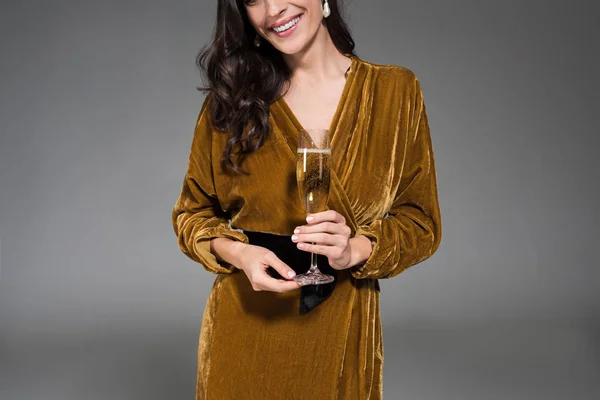 Abgeschnittene Ansicht einer lächelnden Frau in goldenem Kleid, die ein Glas Champagner auf grau isoliert hält — Stockfoto