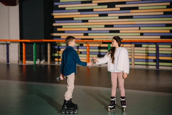 Pretty boy giving to beautiful friend bottle of water, standing on spacious roller rink — Stock Photo
