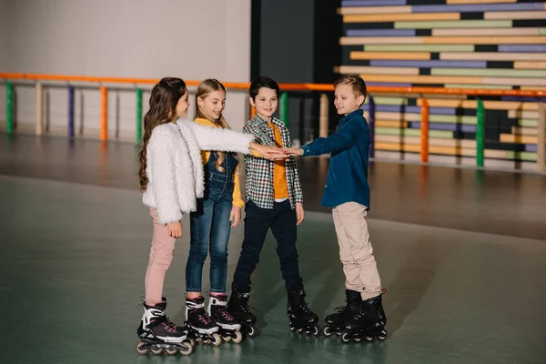 Niños sonrientes en patines de ruedas que se quedan en la pista de patinaje y estiran las manos juntos - foto de stock