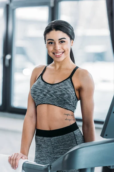 Beautiful sporty african american girl exercising on treadmill and smiling at camera in gym — Stock Photo
