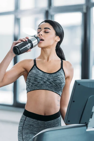 Joven deportivo afroamericano mujer beber agua y hacer ejercicio en la cinta de correr en el gimnasio - foto de stock