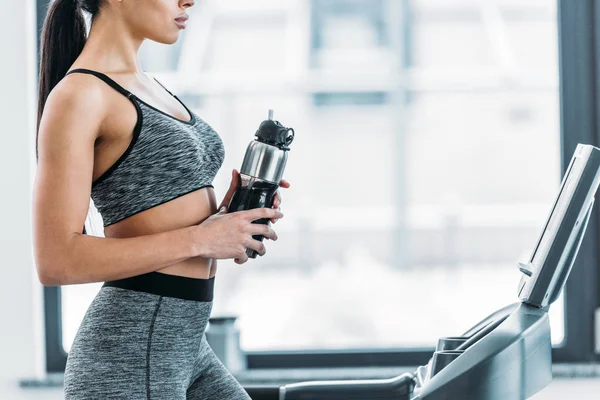 Ritagliato colpo di sporty ragazza afro-americana in possesso di bottiglia sportiva e la formazione sul tapis roulant in palestra — Foto stock