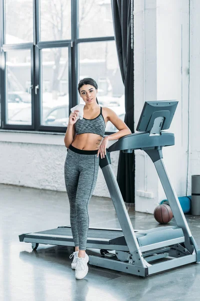 Schöne sportliche afrikanisch-amerikanische Mädchen mit Handtuch lehnt am Laufband und schaut in die Kamera in der Turnhalle — Stockfoto