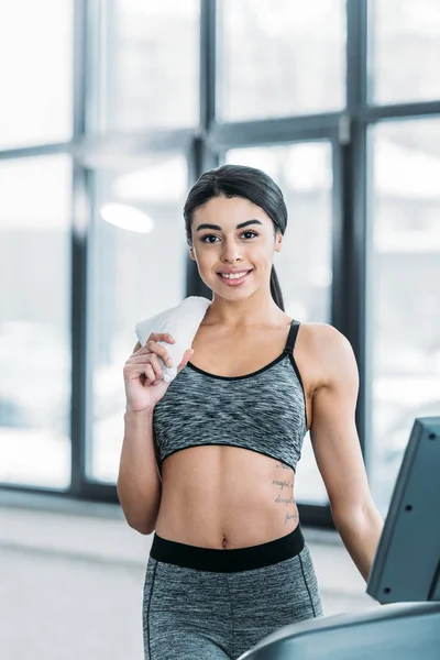 Beautiful sporty african american girl in sportswear holding towel and smiling at camera in gym — Stock Photo