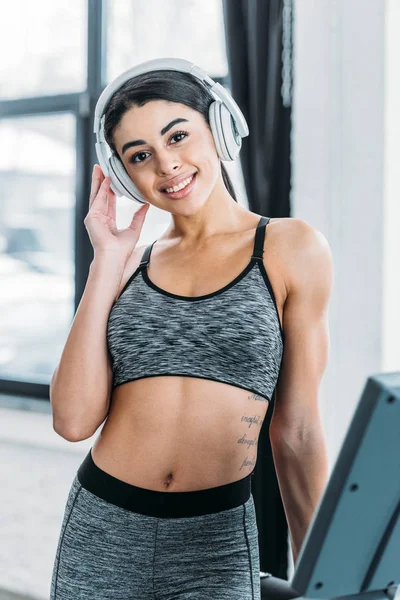 Beautiful sporty african american girl in headphones smiling at camera in gym — Stock Photo