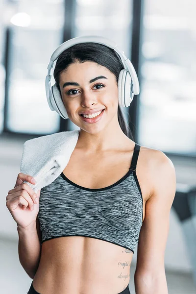 Atractiva joven afroamericana deportista en auriculares sosteniendo la toalla y sonriendo a la cámara en el gimnasio - foto de stock