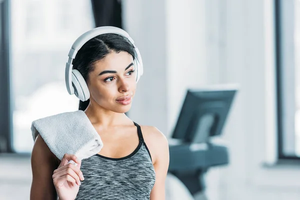 Schöne afrikanisch-amerikanische Sportlerin mit Kopfhörer, Handtuch haltend und im Fitnessstudio wegschauend — Stockfoto