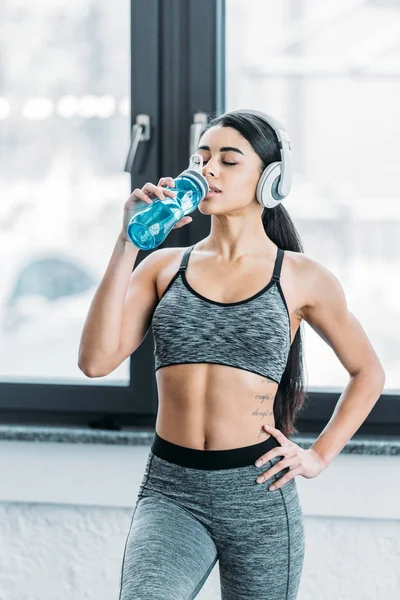 African american sportswoman in headphones drinking water in gym — Stock Photo