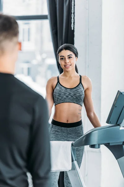 Selective focus of young african american woman standing on treadmill and looking at sporty man on foreground in gym — Stock Photo