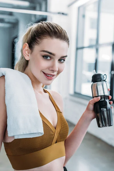 Attractive sporty girl with towel on shoulder holding sports bottle and smiling at camera in gym — Stock Photo