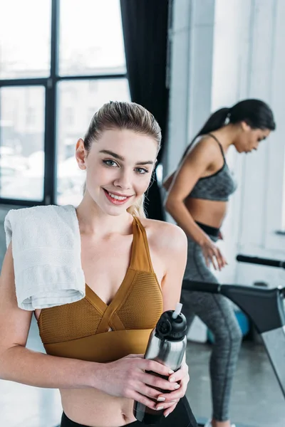 Beautiful young sportswoman with towel and bottle of water smiling at camera, african american girl exercising on treadmill behind in gym — Stock Photo