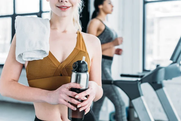 Cropped shot of smiling sporty girl with towel on shoulder holding sports bottle in gym — Stock Photo