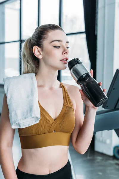 Sporty young woman with towel on shoulder drinking water from sports bottle in gym — Stock Photo
