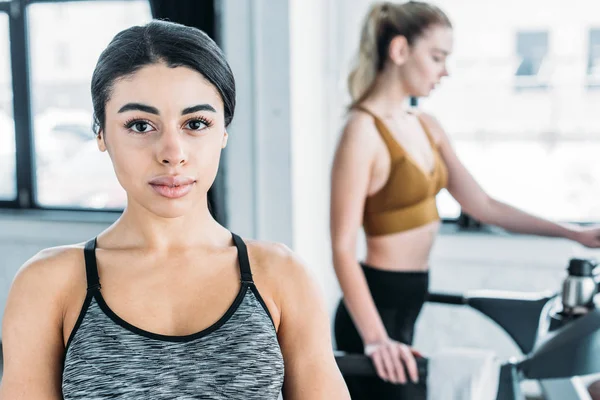 Beautiful african american sportswoman looking at camera and sporty girl exercising on treadmill behind in gym — Stock Photo
