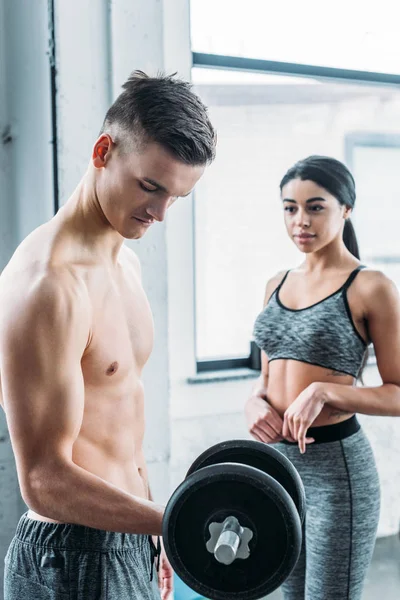 Young african american woman looking at shirtless muscular man training with dumbbell in gym — Stock Photo