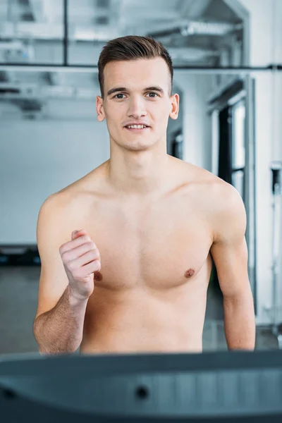 Handsome shirtless sportsman training on treadmill and smiling at camera in gym — Stock Photo