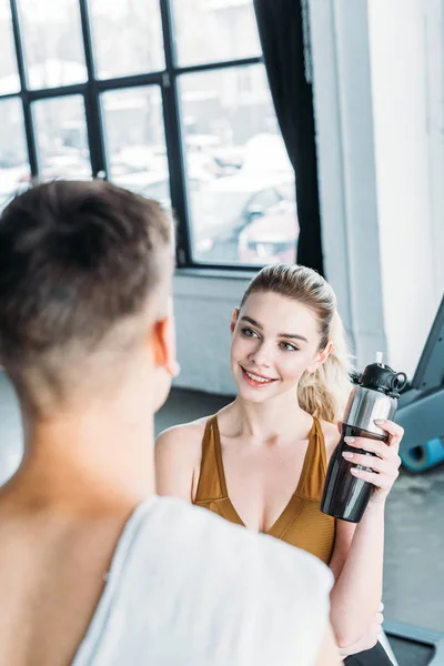 Smiling young woman holding sports bottle and looking at athletic man with towel on foreground in gym — Stock Photo
