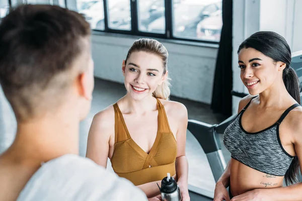 Selective focus of smiling sporty multiethnic girls looking at young sportsman in gym — Stock Photo