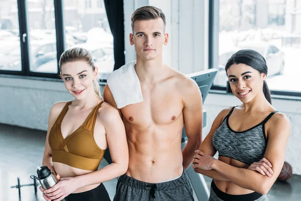 Shirtless male trainer and beautiful multiethnic sportswomen smiling at camera in gym — Stock Photo