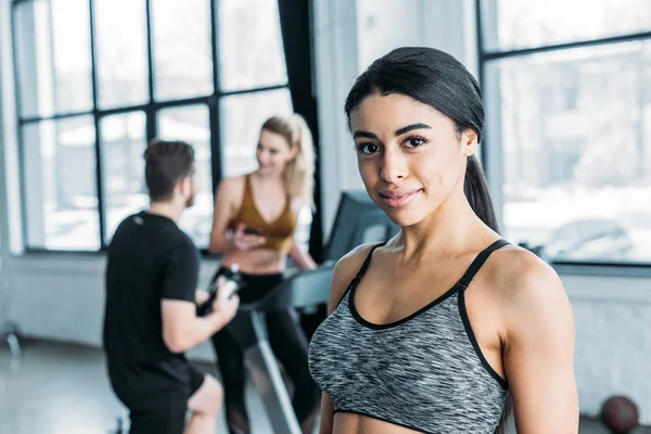 Beautiful african american sportswoman smiling at camera, young friends training behind in gym — Stock Photo