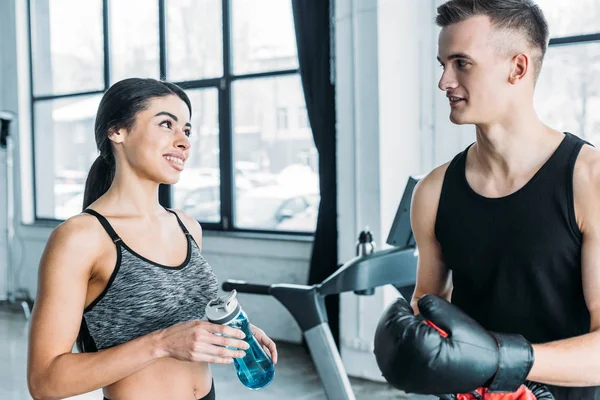 Jeune homme avec des gants de boxe et femme afro-américaine avec bouteille de sport se souriant dans la salle de gym — Photo de stock