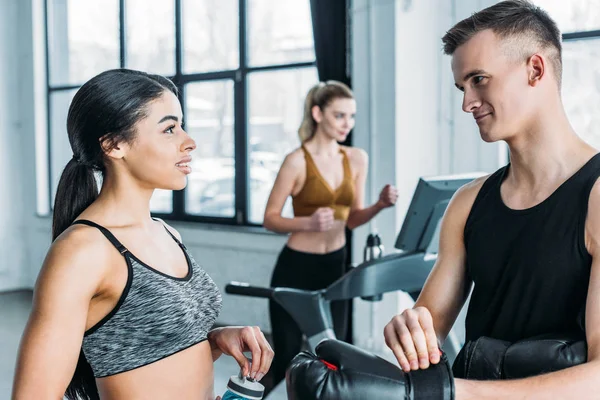Sportlicher junger Mann und Frau, die sich im Fitnessstudio unterhalten und anschauen, Sportlerin beim Training auf dem Laufband hinter sich — Stockfoto