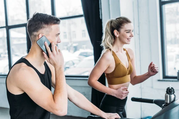 Uomo sorridente parlando da smartphone e guardando felice ragazza sportiva in esecuzione sul tapis roulant in palestra — Foto stock