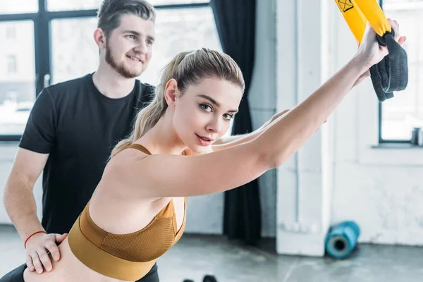 Trainer helping attractive sporty girl exercising with resistance bands and smiling at camera in gym — Stock Photo