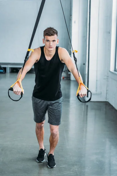 Ganzkörperansicht eines gut aussehenden, muskulösen jungen Mannes, der mit Widerstandsbändern trainiert und im Fitnessstudio in die Kamera schaut — Stockfoto