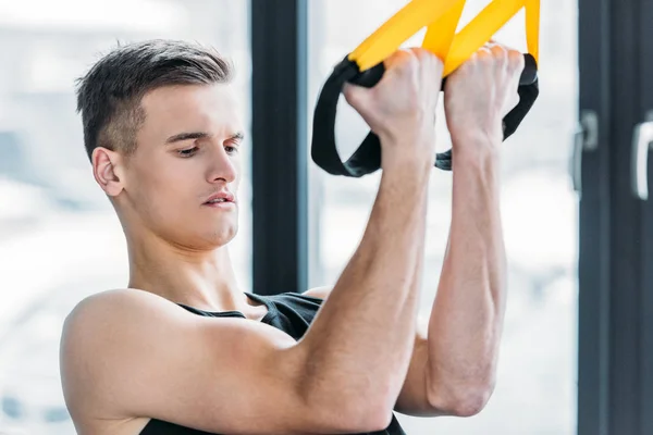 Handsome muscular young sportsman training with suspension straps in gym — Stock Photo